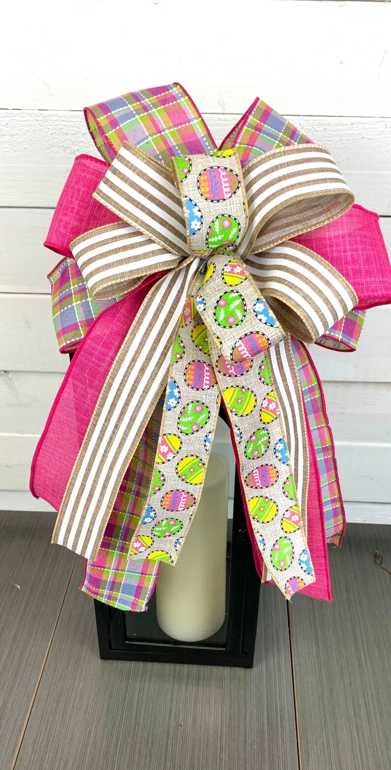 Op de afbeelding: Een roze, wit en groen geruit lint, een bruin en wit gestreept lint en een beige lint met een kleurrijk paasei-patroon zijn gebonden tot een strik om een ​​zwarte lantaarn.
