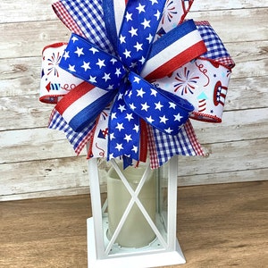 May include: A white lantern with a red, white and blue bow on top. The bow is made of various patterned ribbons, including stars, gingham and fireworks.
