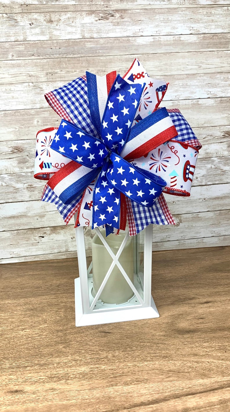 May include: A white lantern with a red, white and blue bow on top. The bow is made of various patterned ribbons, including stars, gingham and fireworks.