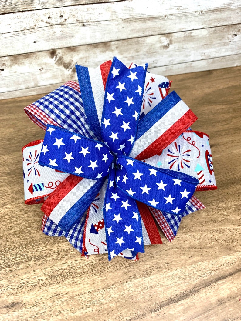 May include: A red, white and blue bow made with various patterned ribbons, including gingham, stars and fireworks. The bow is perfect for a patriotic celebration.