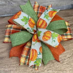 K&ouml;nnte beinhalten: Ein Herbst-Schleife mit verschiedenen B&auml;ndern, darunter ein gr&uuml;nes Leinenband, ein kariertes Band in Orange- und Braunt&ouml;nen und ein wei&szlig;es Band mit einem K&uuml;rbisdruck. Die Schleife ist zu einem dekorativen Knoten gebunden und hat ein rustikales, l&auml;ndliches Flair.