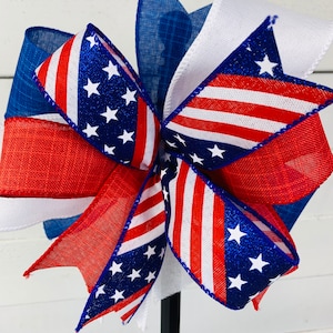 May include: A red, white, and blue patriotic bow with glitter stars. The bow is made of burlap and ribbon and is tied with a wire.