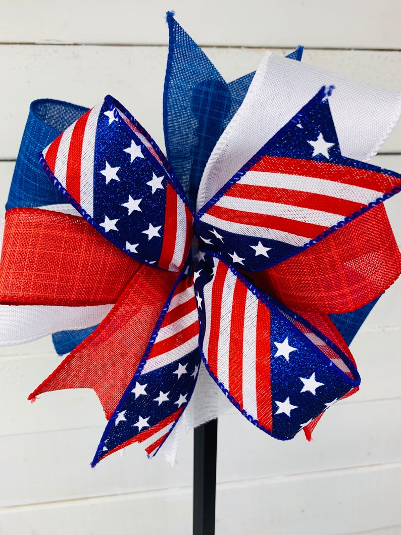 May include: A red, white, and blue patriotic bow with glitter stars. The bow is made of burlap and ribbon and is tied with a wire.