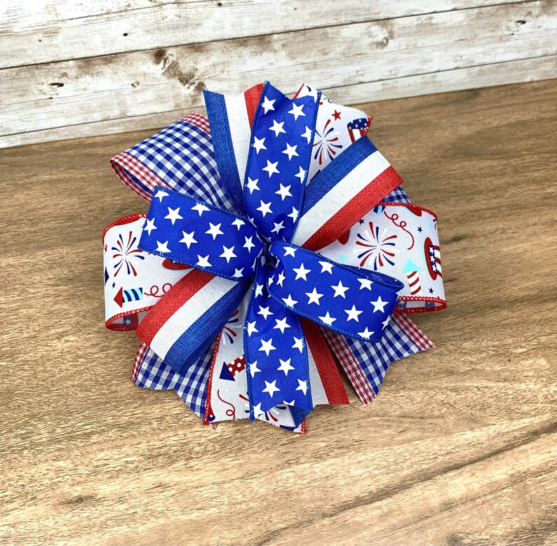 May include: A red, white and blue bow with a gingham pattern, stars and fireworks. The bow is made of ribbon and is perfect for a patriotic celebration.