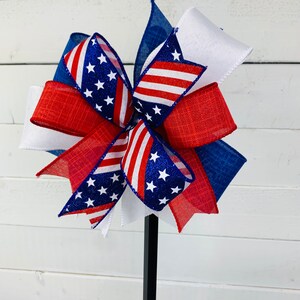 May include: A red, white, and blue bow with a patriotic theme. The bow is made of ribbon with stars and stripes. The bow is attached to a black stick.