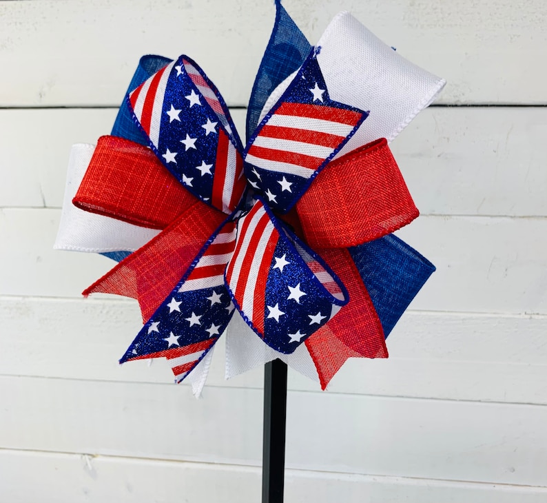 May include: A red, white, and blue bow with a patriotic theme. The bow is made of ribbon with stars and stripes. The bow is attached to a black stick.