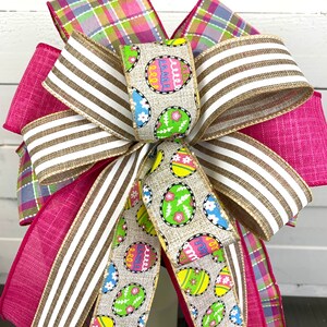 Op de afbeelding: Een roze, witte en groene paas strik met een jute lint met een patroon van kleurrijke eieren. De strik is gebonden met een geruit lint en een gestreept lint.
