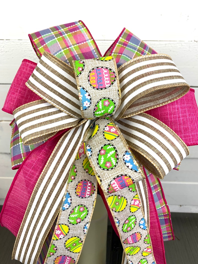 Op de afbeelding: Een roze, witte en groene paas strik met een jute lint met een patroon van kleurrijke eieren. De strik is gebonden met een geruit lint en een gestreept lint.