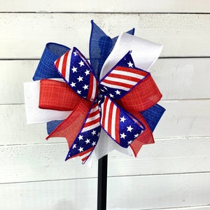 May include: A red, white, and blue bow with a star pattern. The bow is made of burlap and ribbon and is tied with a wire.
