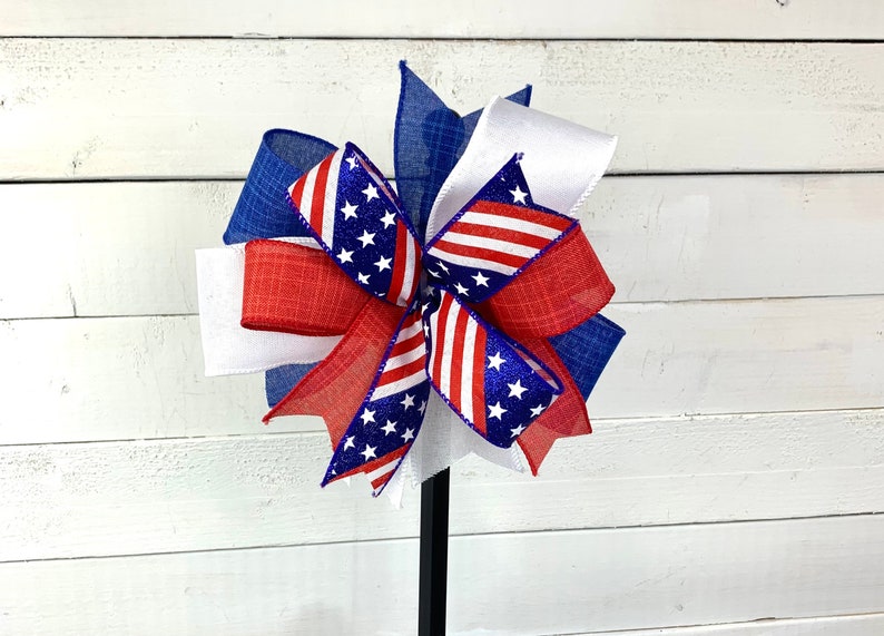 May include: A red, white, and blue bow with a star pattern. The bow is made of burlap and ribbon and is tied with a wire.