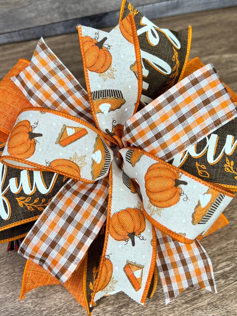 Op de afbeelding: Een herfstthema strik met oranje, bruin en wit geruit lint, een bruin lint met het woord "dankbaar" in wit, en een wit lint met oranje pompoenen en taartontwerpen.