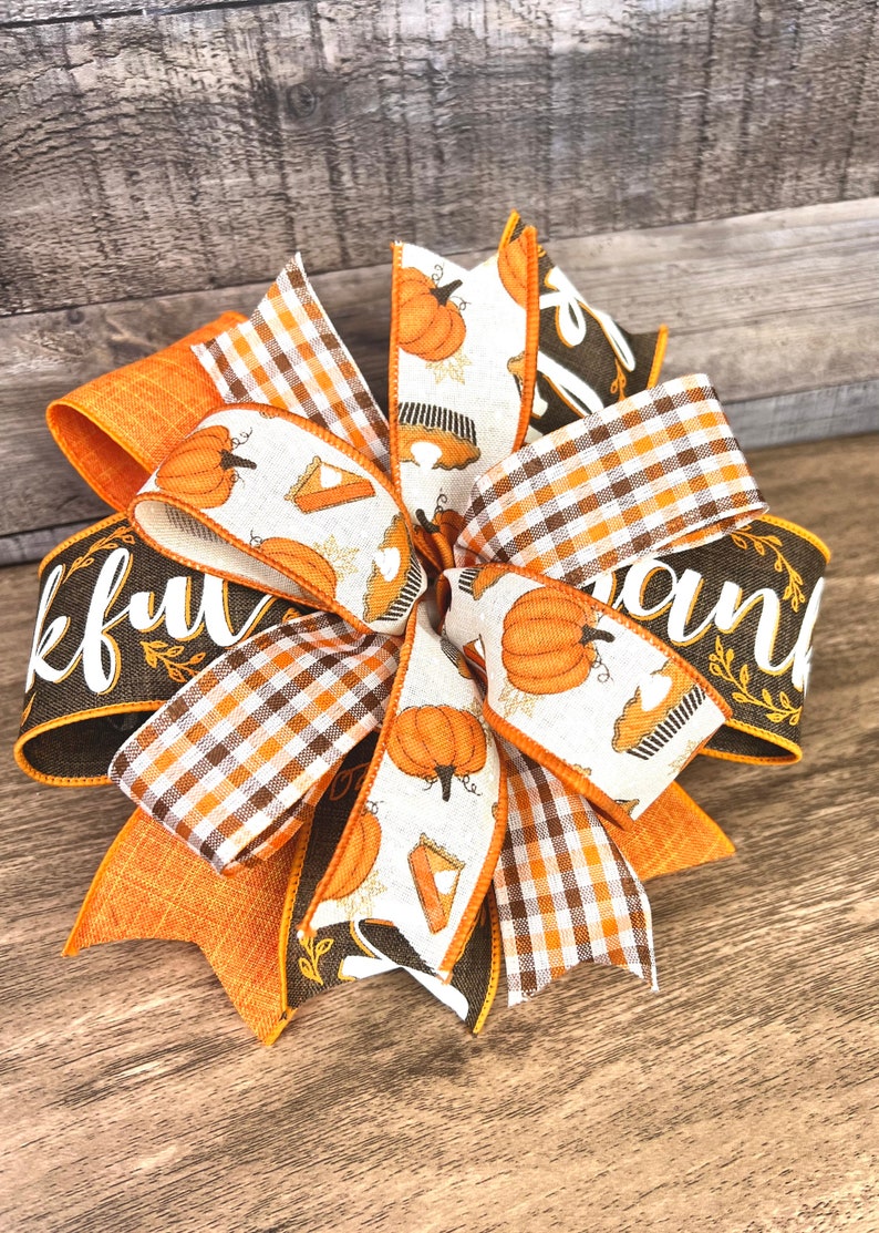 Op de afbeelding: Een Thanksgiving-strik met oranje, bruine en witte linten. De linten hebben een geruit patroon, een pompoen- en taartontwerp en de woorden "Thankful" en "and".