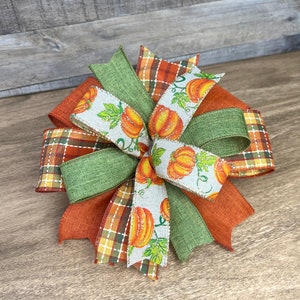 K&ouml;nnte beinhalten: Eine herbstliche Schleife mit orangefarbenem, gr&uuml;nem und wei&szlig;em Band. Das Band hat ein Karomuster, eine einfarbige gr&uuml;ne Farbe und ein wei&szlig;es Band mit einem K&uuml;rbisdruck.