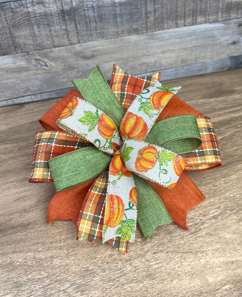 K&ouml;nnte beinhalten: Eine herbstliche Schleife mit orangefarbenem, gr&uuml;nem und wei&szlig;em Band. Das Band hat ein Karomuster, eine einfarbige gr&uuml;ne Farbe und ein wei&szlig;es Band mit einem K&uuml;rbisdruck.