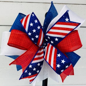 May include: A red, white, and blue bow with a glittery American flag design. The bow is made of multiple layers of ribbon and is tied with a knot in the center.