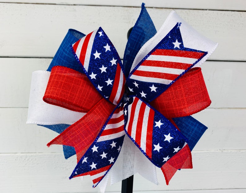 May include: A red, white, and blue bow with a glittery American flag design. The bow is made of multiple layers of ribbon and is tied with a knot in the center.
