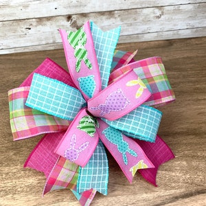 May include: A pink, blue, and green plaid Easter bow with bunny and egg designs. The bow is made of ribbon and is tied in a knot.