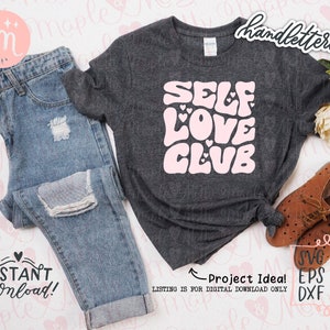 May include: A gray t-shirt with the text "SELF LOVE CLUB" in pink with hearts. The text is in a retro style font. The t-shirt is on a white surface with a pair of jeans, a pair of brown shoes, and flowers.