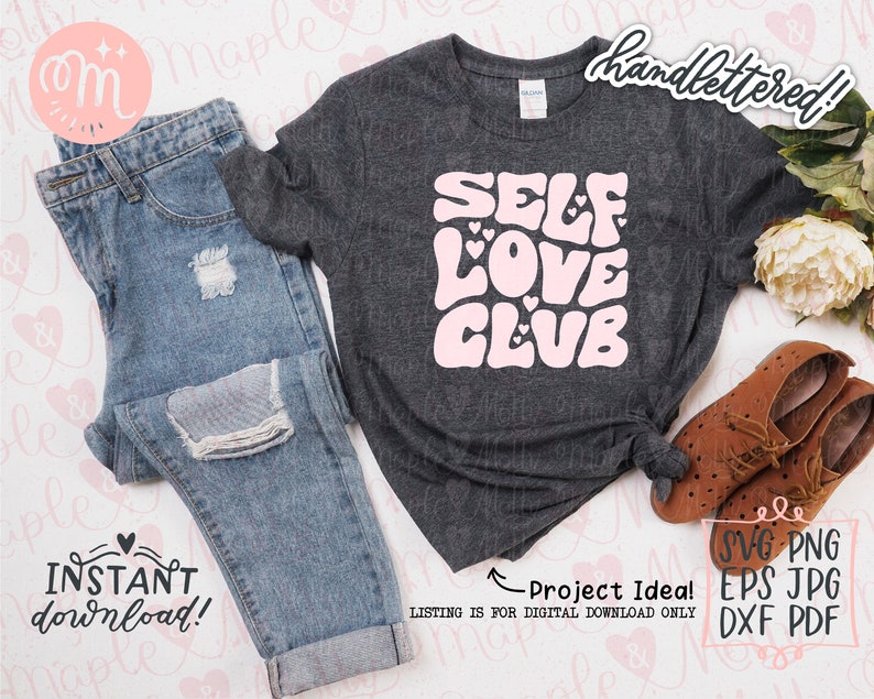 May include: A gray t-shirt with the text "SELF LOVE CLUB" in pink with hearts. The text is in a retro style font. The t-shirt is on a white surface with a pair of jeans, a pair of brown shoes, and flowers.