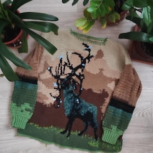 Peut inclure: Un pull crocheté vert, marron et beige avec un motif de cerf. Le cerf est marron avec un corps vert et a de grands bois.