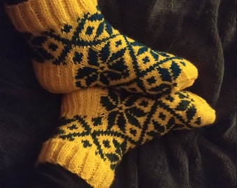 May include: A pair of yellow and green knitted socks with a geometric pattern. The socks are folded over and are sitting on a gray fuzzy surface.