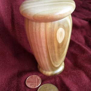 May include: A wooden box with a lid, made of light brown wood with a striped pattern. The box is sitting on a red surface with two British coins.