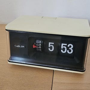 May include: A vintage Caslon flip clock displaying 5:53. The clock has a cream-colored top and a black face with white numerals. The clock is rectangular with a clear plastic front and sides. The brand name "CASLON" is visible.