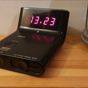 May include: A black alarm clock with a digital display showing the time 13.23. The clock has a variety of buttons and controls.