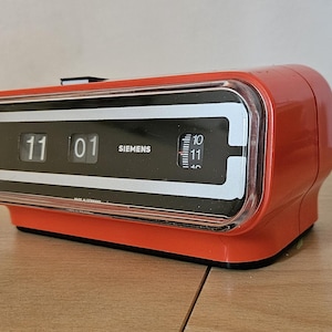 May include: A red vintage flip clock alarm clock with a clear plastic cover. The clock displays the time 11:01 and has the word "SIEMENS" printed on the face.