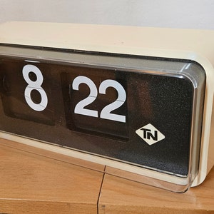 Könnte beinhalten: Eine weiße Flip-Uhr mit schwarzen Zahlen, die 8:22 anzeigen. Die Uhr hat eine transparente Plastikabdeckung und einen schwarzen Hintergrund mit den Buchstaben "TN" in einer Raute.