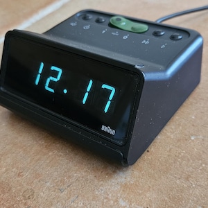 Puede incluir: Un reloj despertador digital negro que muestra la hora 12:17 en dígitos azules. El reloj tiene varios botones en la parte superior y la marca "Braun" es visible. El reloj está enchufado.