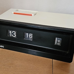 Könnte beinhalten: Eine weiße und schwarze Flip-Uhr mit der Zeit 13:16. Die Uhr hat ein schwarzes Zifferblatt mit weißen Zahlen und einen schwarzen Rahmen. Die Uhr wird von AEG hergestellt.