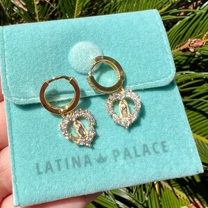 Puede incluir: Pendientes de aro dorados con colgantes en forma de coraz&oacute;n. Cada colgante presenta una figura religiosa central rodeada de cristales transparentes. Los pendientes se muestran en una bolsa de terciopelo azul claro con el texto "LATINA PALACE".
