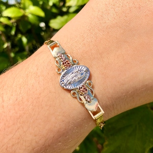 Może przedstawiać: Złota bransoletka z zapięciem w kształcie serca i zawieszką przedstawiającą wizerunek religijny. Zawieszka jest otoczona małymi sercami i różami.