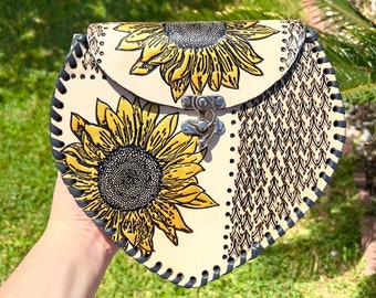 May include: A heart-shaped leather purse with a black stitched border. The purse features a sunflower design in yellow and black on a cream background.