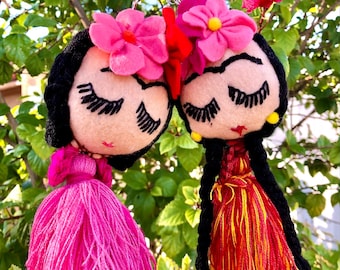 May include: Two colorful felt doll keychains with black hair, pink and red dresses, and floral headbands. The dolls have embroidered eyelashes and are looking down.