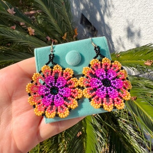 K&ouml;nnte beinhalten: Ein Paar Ohrringe in Blumenform, die aus rosa, lila, gelben und schwarzen Perlen gefertigt sind. Die Ohrringe werden auf einem t&uuml;rkisfarbenen Samtbeutel pr&auml;sentiert.