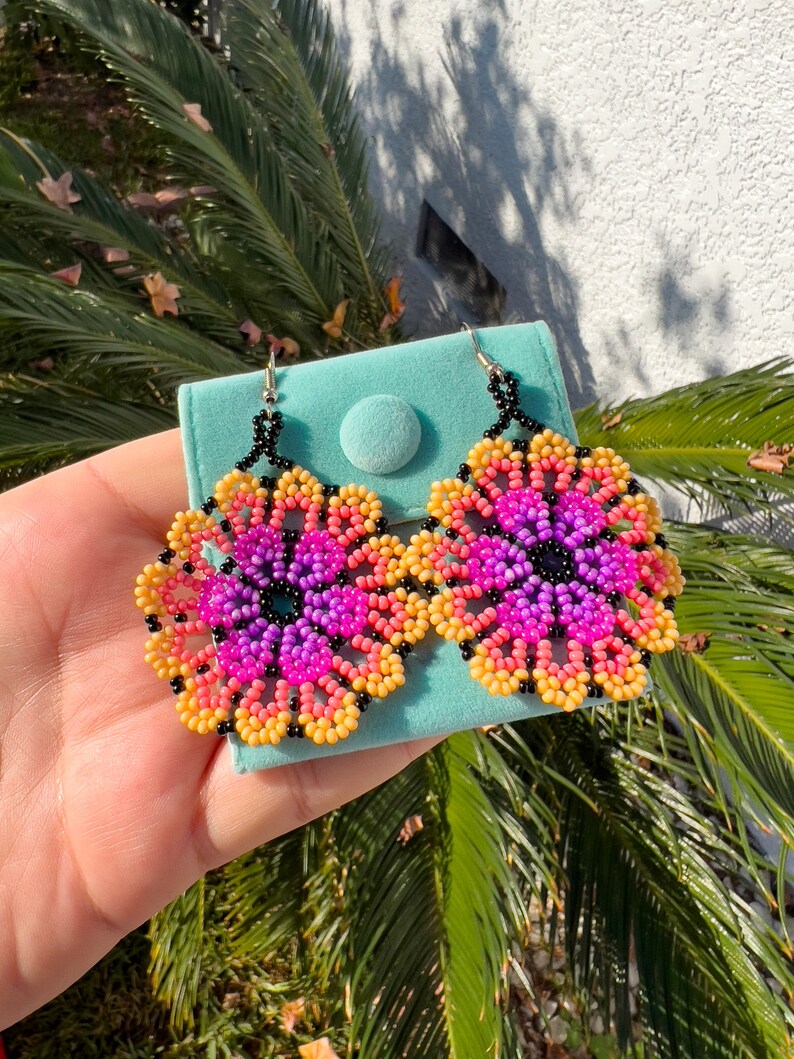 K&ouml;nnte beinhalten: Ein Paar Ohrringe in Blumenform, die aus rosa, lila, gelben und schwarzen Perlen gefertigt sind. Die Ohrringe werden auf einem t&uuml;rkisfarbenen Samtbeutel pr&auml;sentiert.