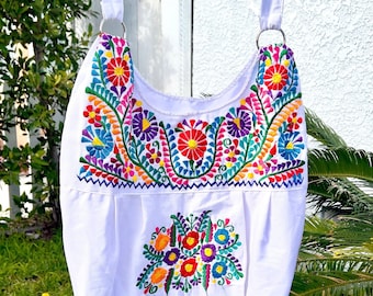 May include: White canvas tote bag with colorful embroidered flowers and leaves. The bag has a long white strap and a silver ring on each side.