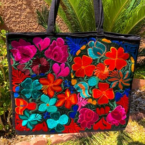 May include: Black tote bag with colorful embroidered flowers in shades of pink, red, orange, yellow, blue, and green. The bag has two black straps.