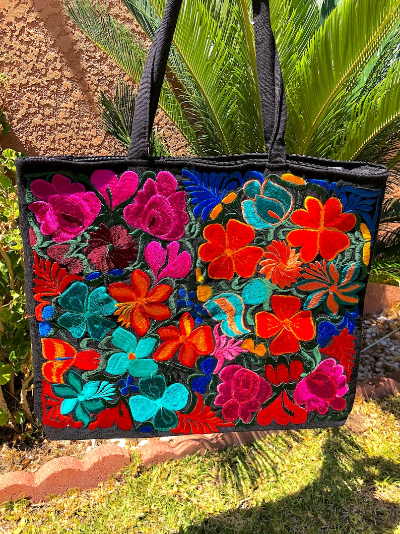 May include: Black tote bag with colorful embroidered flowers in shades of pink, red, orange, yellow, blue, and green. The bag has two black straps.