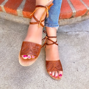 May include: Brown leather sandals with intricate floral carvings. The sandals have laces that tie up the ankles and feature a light brown colour. The sandals are worn by a person with pink nail polish.