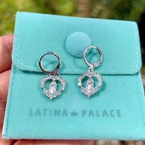 Puede incluir: Pendientes de aro plateados con colgantes en forma de coraz&oacute;n. Cada colgante presenta una figura religiosa rodeada de piedras transparentes. Los pendientes se exhiben en una bolsa de terciopelo turquesa con el texto "LATINA PALACE".