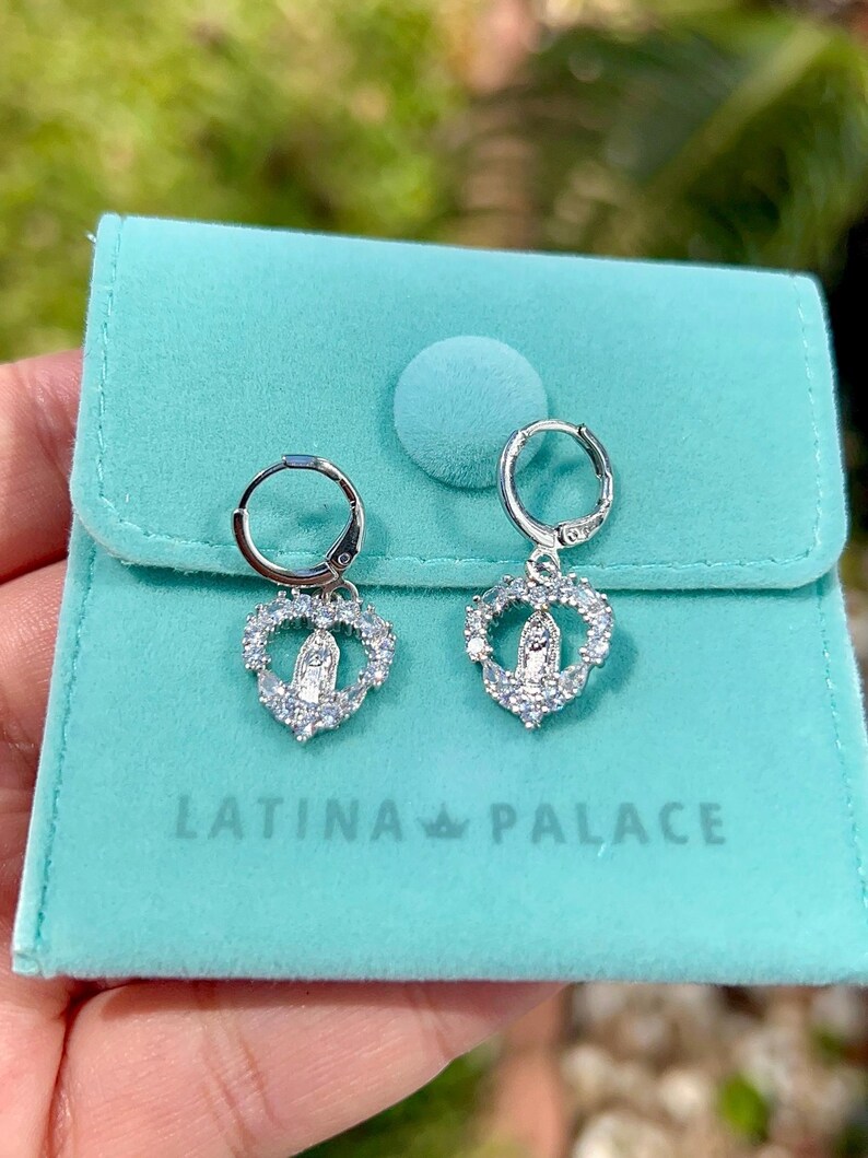 Puede incluir: Pendientes de aro plateados con colgantes en forma de coraz&oacute;n. Cada colgante presenta una figura religiosa rodeada de piedras transparentes. Los pendientes se exhiben en una bolsa de terciopelo turquesa con el texto "LATINA PALACE".