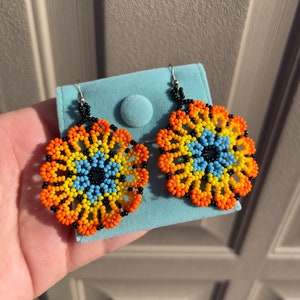 K&ouml;nnte beinhalten: Ein Paar Perlenohrringe mit einem runden Design. Die Ohrringe sind aus schwarzen, orangefarbenen, gelben und blauen Perlen gefertigt. Die Ohrringe h&auml;ngen an silbernen Haken.