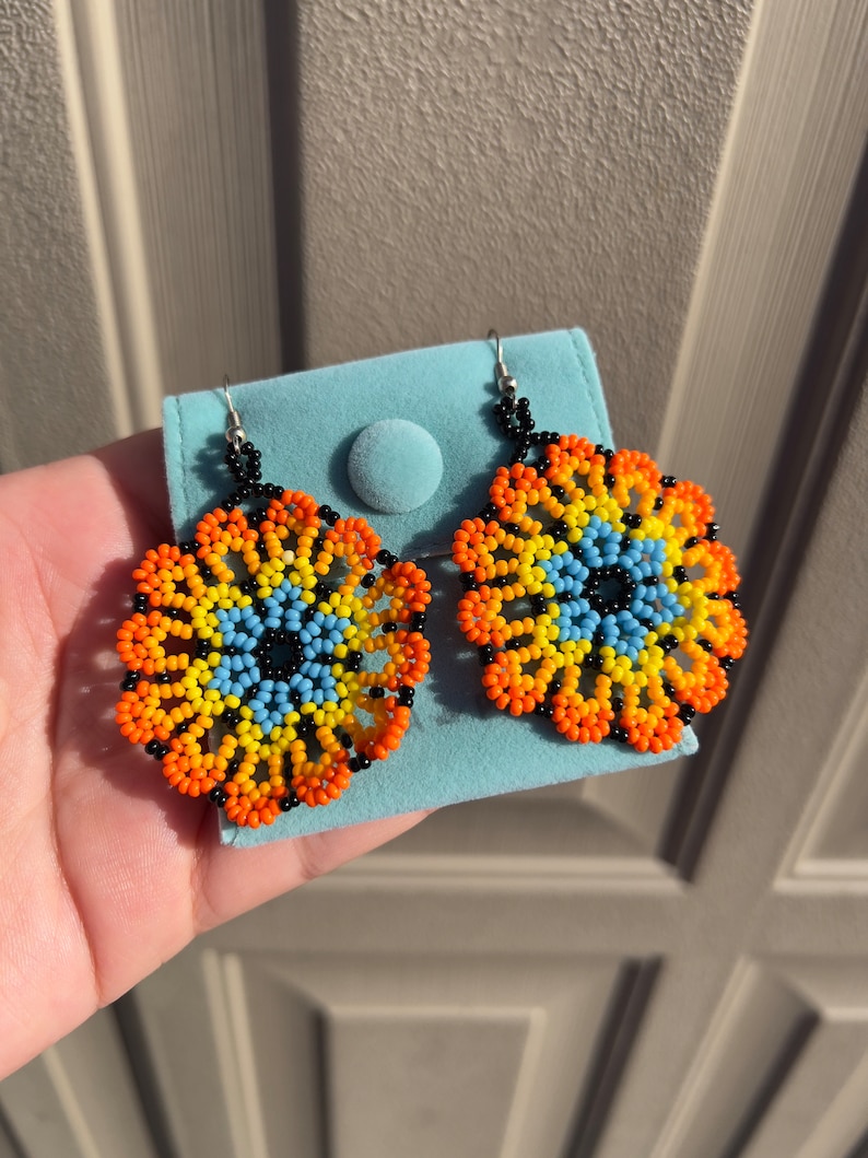 K&ouml;nnte beinhalten: Ein Paar Perlenohrringe mit einem runden Design. Die Ohrringe sind aus schwarzen, orangefarbenen, gelben und blauen Perlen gefertigt. Die Ohrringe h&auml;ngen an silbernen Haken.