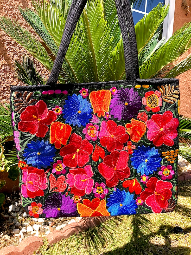 May include: Black tote bag with colorful embroidered flowers in shades of red, pink, orange, purple, blue, and yellow. The flowers are arranged in a random pattern on the bag.