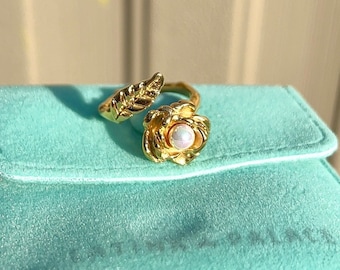 Può includere: Un anello d'oro con un designo a forma di perla e foglia. L'anello è su una scatola di velluto turchese.