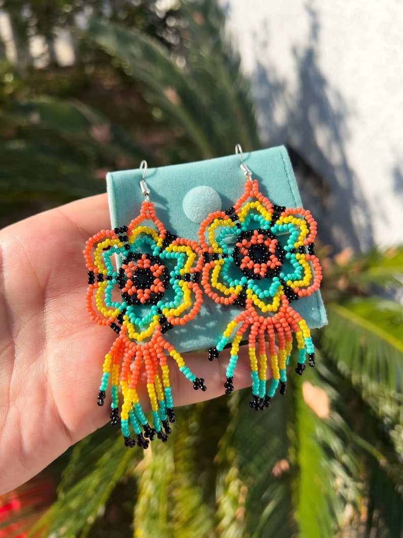 May include: A pair of colorful beaded earrings with a flower design and fringe. The earrings are made with orange, yellow, turquoise, and black beads.