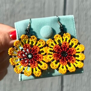 K&ouml;nnte beinhalten: Ein Paar Perlenohrringe mit Blumenmuster. Die Ohrringe sind aus schwarzen, roten, orangefarbenen und gelben Perlen gefertigt. Die Ohrringe befinden sich auf einem hellblauen Hintergrund.